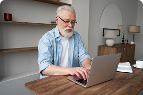 Senior man using a laptop at a wooden table, emphasizing technology use for accessing dental and vision insurance information.