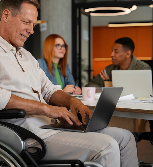Man in a wheelchair using a laptop in a collaborative office setting, emphasizing accessibility and productivity for professionals.