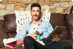 Man sitting on a couch with a small dog, holding a tablet, surrounded by cozy home decor, illustrating the comfort and companionship of pet ownership in the context of pet insurance.