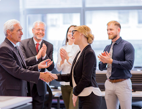 Business professionals shaking hands in a modern office setting, celebrating a successful partnership or agreement, with colleagues applauding in the background.