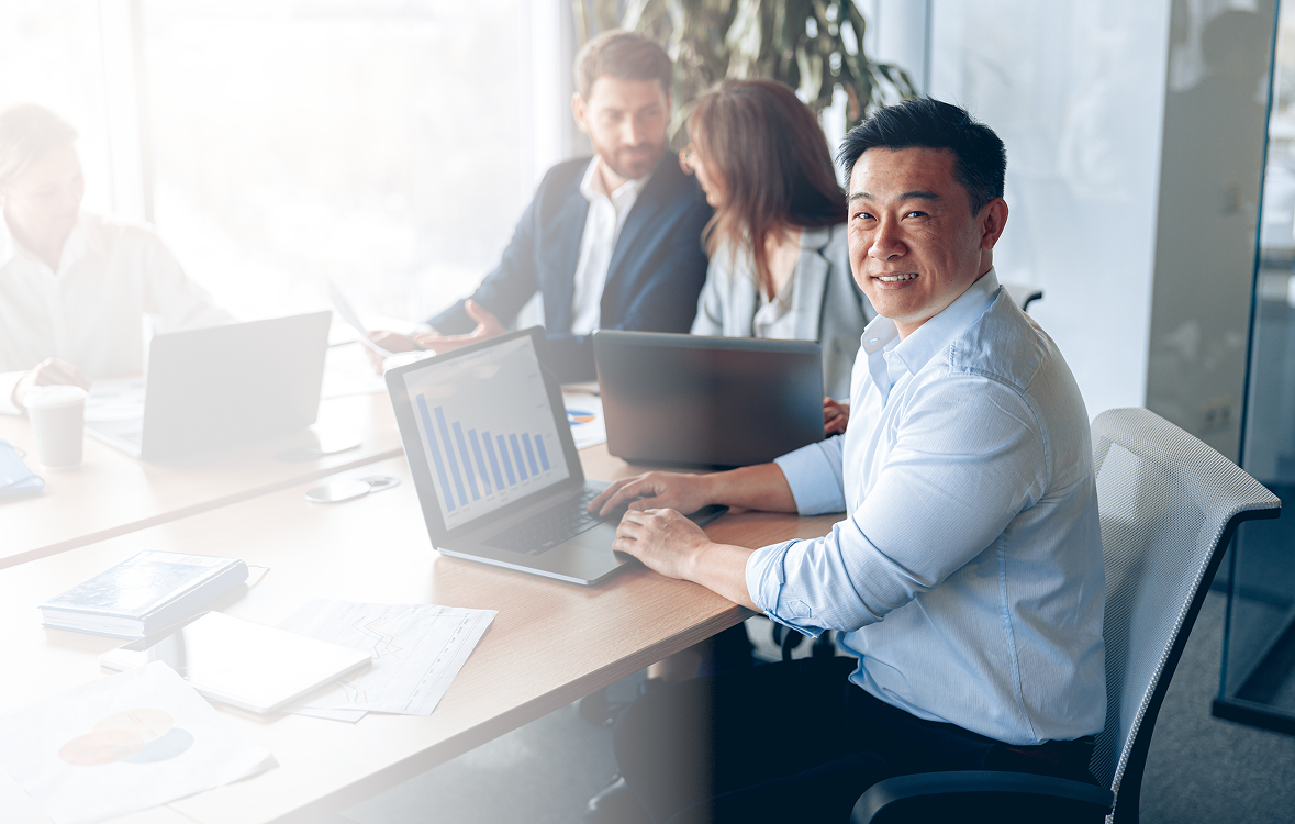 Business professional smiling at a laptop in a meeting room, surrounded by colleagues discussing charts and data, emphasizing teamwork and collaboration for business growth.