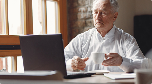 Elderly man sitting at a table with a laptop, holding a cup, contemplating financial security and final expense insurance options.