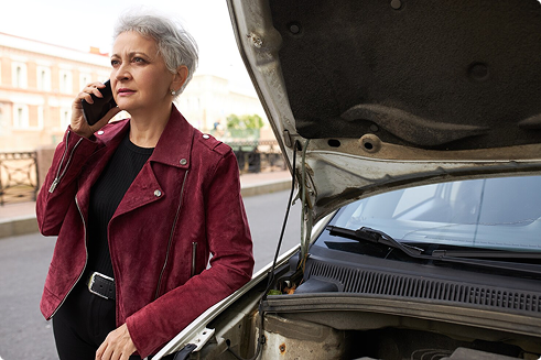 Senior woman on phone beside a car with open hood, reflecting the importance of roadside assistance in senior auto insurance coverage options.
