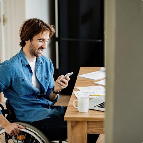 Man in a wheelchair smiling while using a smartphone at a desk with a laptop and documents, representing self-employed professionals considering simplified issue disability coverage.