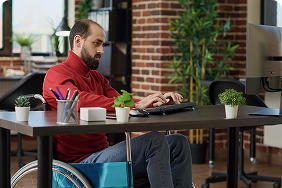 Man in a wheelchair working on a laptop at a modern office desk, emphasizing accessibility and professionalism in the context of disability coverage for self-employed individuals and business owners.