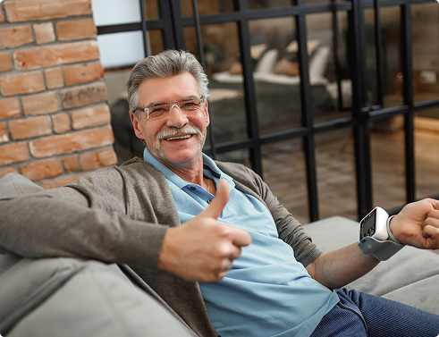 Smiling older man giving a thumbs-up while sitting on a couch, showcasing a positive attitude in a cozy, modern living space.
